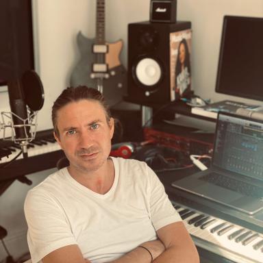 Man with brown hair tied in pony tail wearing white t-shirt sits with arms crossed in chair next to keyboard